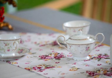 Bir çaydanlık ve iki fincan çiçekli masa örtüsü olan bir masa. Çaydanlık bir fincan tabağında, bardaklar da tepside. Masa örtüsünün çiçek deseni var ve fincanlar ve çay tabağı beyaz.