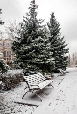 A bench is sitting in the snow next to two trees. The bench is empty and covered in snow. The trees are bare and covered in snow, giving the scene a peaceful and serene atmosphere