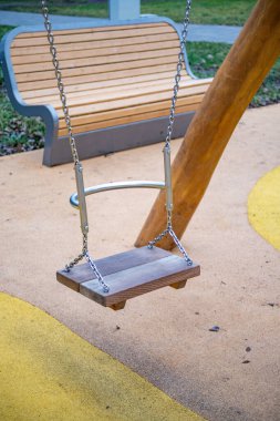 A swing is suspended from a wooden post. The swing is empty. A bench is in the background