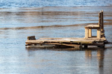 Suda tahta bir bank yüzüyor. Bankta kırık ve sandalyeye bağlı bir sandalye var. Su sakin ve gökyüzü berrak.