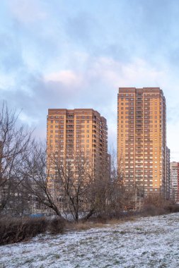 Önünde iki yüksek bina ve bir ağaç olan bir şehir silueti. Gökyüzü bulutlu ve güneş batıyor.