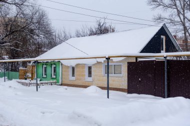Yeşil çatılı bir ev karla kaplıdır. Evin etrafı çitlerle çevrili ve kardan çıkmış metal bir direği var. Kar yere yığıldı ve gökyüzü bulutlu.