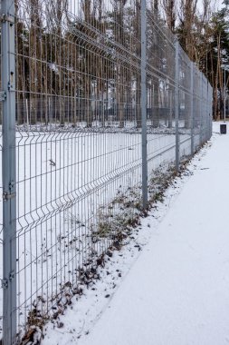 Üzerinde tel örgü ve kar olan bir çit. Çit ağaçlar ve çalılarla çevrili.