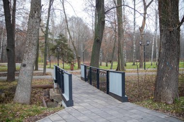 Arka planda ağaçlar olan bir parktaki derenin üzerindeki köprü. Köprü metalden yapılmıştır ve siyah beyaz bir tasarımı vardır.