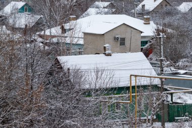 Çatıdan sarkan bacası ve borusu olan karlı bir ev. Ev ağaçlarla çevrili ve karlar yere yığılmış.