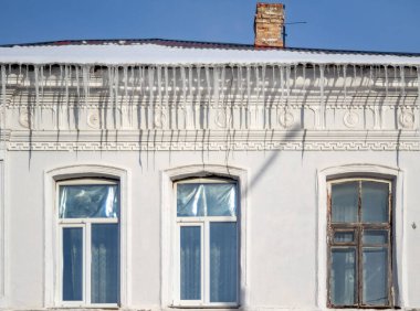 Çatıdan sarkıtlar sarkan beyaz bir bina. Binanın iki penceresi ve bir bacası var.