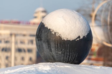 Karla kaplı büyük siyah beyaz bir top. Kar topun üzerine yığılmış, bu da onu dev bir kartopu gibi gösteriyor. Sahne bir binanın arka planında geçiyor.