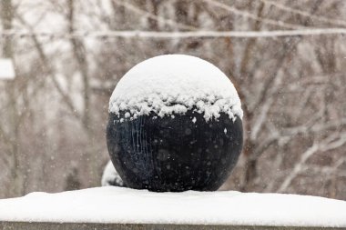 Büyük siyah bir top karla kaplıdır. Kar topun üzerine yığılmış, bu da onu dev bir kartopu gibi gösteriyor. Sahne, arka planda ağaçlarla karlı bir ortamda geçiyor.