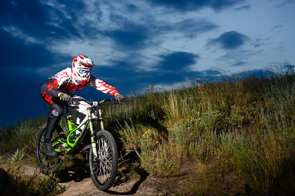 Ciclista de downhill profissional totalmente equipado montando a bicicleta na trilha rochosa noturna — Fotografia de Stock