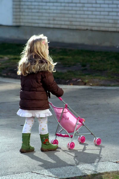 The girl rides her doll in a stroller, has green boots and a brown winter jacket the stroller is pink