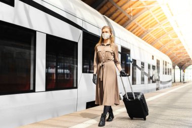 Koruyucu maskeli ve eldivenli genç bir kadın. Toplu taşıma istasyonunda valiziyle birlikte.