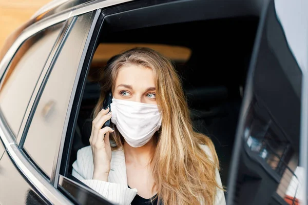 Koruyucu maskeli genç bir iş kadını telefonla konuşuyor ve salgın halinde araba sürüyor. 