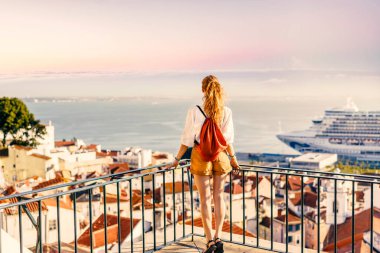 Sarışın bir kadın balkonda duruyor ve gün batımında deniz manzarasına bakıyor. Şapka, mantar çanta, safari şortu ve beyaz gömlek giyiyor.