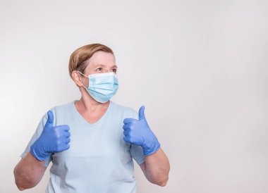 Maske takan ve baş parmağını gösteren yaşlı bir kadın.