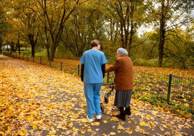 Yürüyen yaşlı bir kadın, mavi üniformalı bir hemşire ya da bakıcının yardımıyla düşmüş yapraklarla parkta yürüyor.