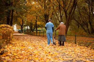 Yürüyen yaşlı bir kadın, mavi üniformalı bir hemşire ya da bakıcının yardımıyla düşmüş yapraklarla parkta yürüyor.