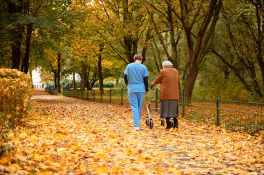 Yürüyen yaşlı bir kadın, mavi üniformalı bir hemşire ya da bakıcının yardımıyla düşmüş yapraklarla parkta yürüyor.