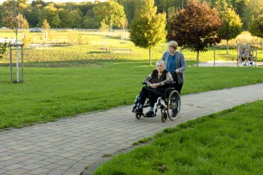 Mavi üniformalı bekçi yaşlı bir kadın hastayla tekerlekli sandalyenin yardımıyla parkta yürüyüş yapıyor. 
