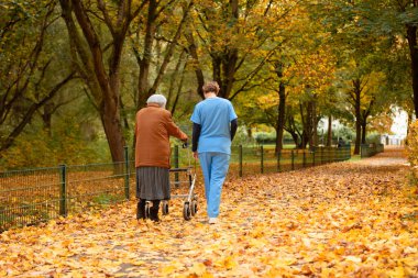 Yürüyen yaşlı bir kadın, mavi üniformalı bir hemşire ya da bakıcının yardımıyla düşmüş yapraklarla parkta yürüyor.