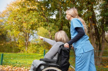 Parkta mavi üniformalı, tekerlekli sandalyede gülümseyen yaşlı, mutlu bir kadın.