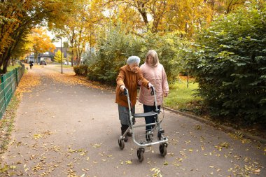 Kadın ve yaşlı annesi, sonbahar yapraklarıyla kaplı parkta yürüyüş yapıyor, aile zamanlarını birlikte geçiriyorlar.