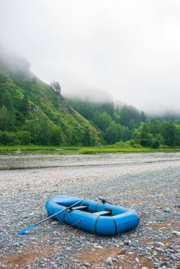Sabahın erken saatlerinde nehir kıyısında bir dağ manzarasının arka planında şişirilebilir mavi bir tekne. Dağlar, nehir, kumsal ve çam ormanı.