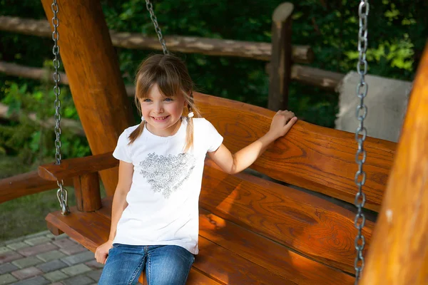 Menina alegre em uma camiseta branca e jeans em um balanço de madeira — Fotografia de Stock