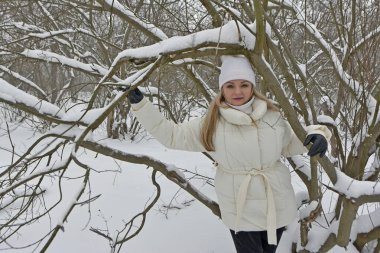 Kışın beyaz ceketli seksi sarışın kadın, beyaz şapka ve siyah kış taytları ormanda bir fotoğraf çekiminde..