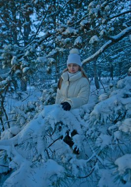 Kışın beyaz ceketli seksi sarışın kadın, beyaz şapka ve siyah kış taytları ormanda bir fotoğraf çekiminde..