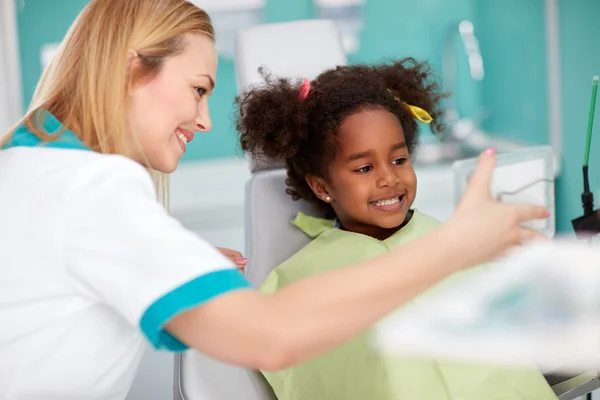 Menina preta agradável na cadeira dental mostrando dentes no espelho para dental — Fotografia de Stock