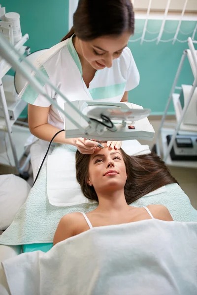 Cosmetologista trata o rosto de uma mulher olhando através de um magni — Fotografia de Stock