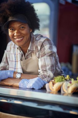 Fast food servisinde çalışan genç bir Afro-Amerikan kadın çalışanın portresi, kameraya bakıyor, gülümsüyor, kıvrak zekalı elleri çapraz, eldiven giyiyor ve sandviçlere gitmeye hazır.. 