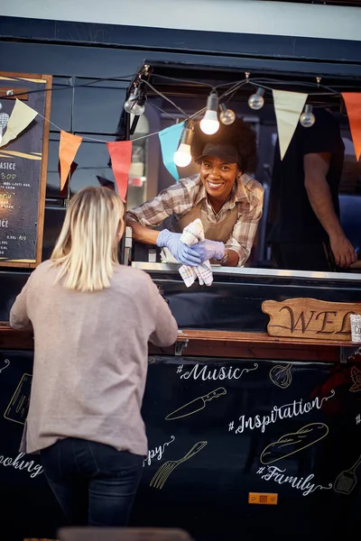 young afro-american female employee talking to a customer from a window of truck modified for fast food, looking at camera with smile. eye contact