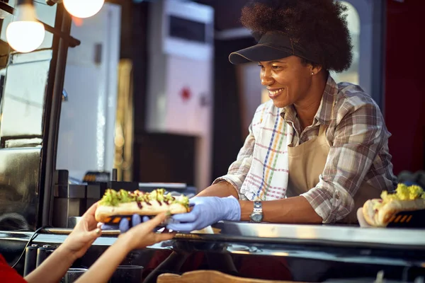 young afro-american female employee in fast food service giving sandwiches to a customer with a smile