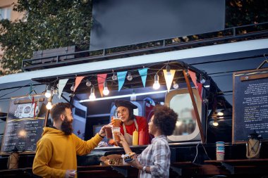 Arkadaşlar fast food servisinin önünde bir çalışanla kadeh kaldırıyor.