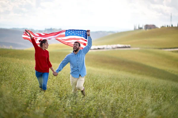 A couple in love having good time running through a meadow on a beautiful sunny day with unfurled American flag above heads. Election, campaign, freedom concept