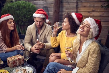 Yeni yıl arifesini kafalarında Noel Baba şapkalarıyla ve ellerinde beyaz şarap bardaklarıyla dışarıda oturarak kutlayan sarhoş bir grup yetişkin.