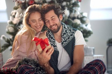 Sakallı, beyaz, gülümseyen, elinde güzel bir sarışınla elinde hediyeyle omuzuna yaslanmış.