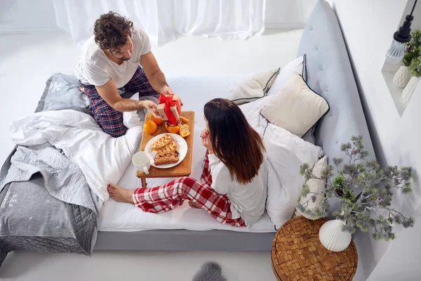 ベッドで朝食をとる若い白人のカップルのトップビュー 男性からの贈り物に驚いた女性は — ストック写真
