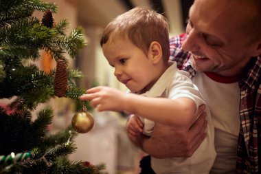Küçük beyaz çocuk Noel ağacına dokunuyor ve babası onu kollarına alıyor.