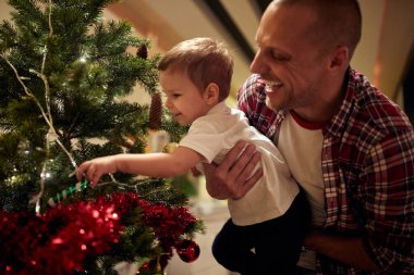 Baba kucağında küçük bir çocuk Noel ağacında süslere dokunuyor, gülüyor, gülümsüyor.