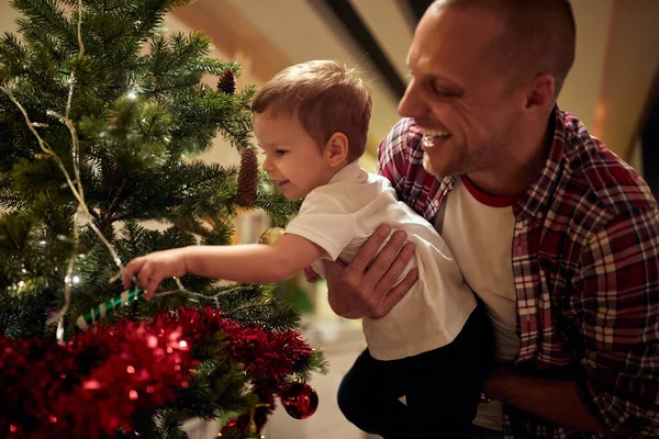 Baba kucağında küçük bir çocuk Noel ağacında süslere dokunuyor, gülüyor, gülümsüyor.