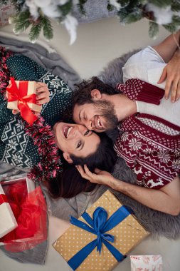 Mutlu, genç, güzel, beyaz bir çiftin Noel 'de yerde yatıp gülmesinin, eğlenmesinin ve etraflarında hediyelerle eğlenmesinin en güzel manzarası. Kadın kameraya bakıyor, göz teması kuruyor.