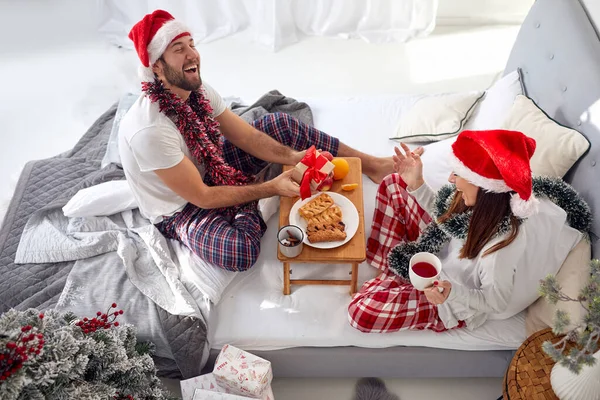 楽しそうに笑顔でベッドで朝食を食べたりクリスマスのプレゼントを交換したりサンタの帽子をかぶったり — ストック写真