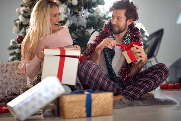 若い白人カップルがクリスマスプレゼントを開けて笑顔で見つめ合いパジャマの床に座り — ストック写真