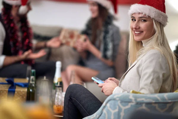 若い白人ブロンドの肖像画サンタの帽子で笑って カメラを見て 携帯電話を持って 背景の贈り物を交換する友人と クリスマスのコンセプト — ストック写真