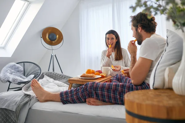 ベッドルームでの朝食中にオレンジを食べることを楽しんでいる若いカップル 一緒に — ストック写真