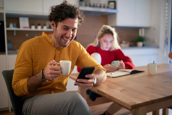 A young couple drinking coffee and surfing the internet on a beautiful morning at the kitchen. Routine, relationship, together