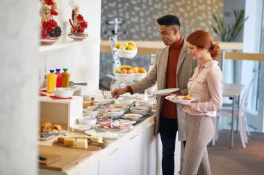 Afro-Amerikalı erkek öğle arasında beyaz kadınla konuşuyor, açık büfeden yemek seçiyor.