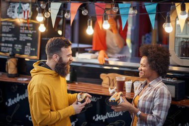 Arkadaşlar fast food servisinin önünde sandviç yerken konuşuyor ve gülüyorlar. Açık hava